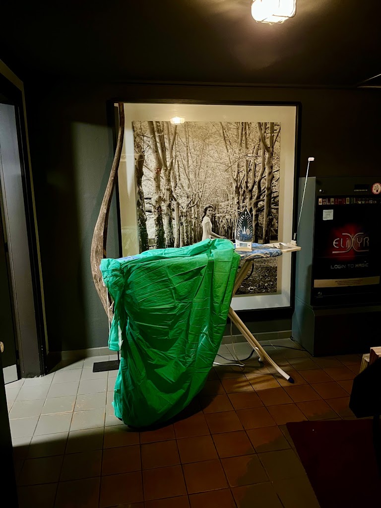 Lit like a renaissance painting, an ironing board is draped in a long green 1790s gown. The ironing board stands in a shaft of life between a bathroom door and a vending machine.