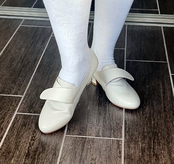 A pair of legs wearing white stockings. On the feet are a pair of white American Duchess Kensington shoes.