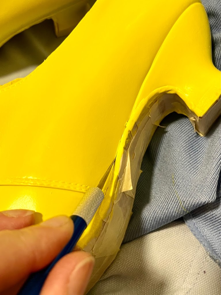 The heel of a bright yellow American Duchess Kensington shoe. A craft knife is cutting away a layer of pale yellow painters tape from the join between leather shoe and leather sole.
