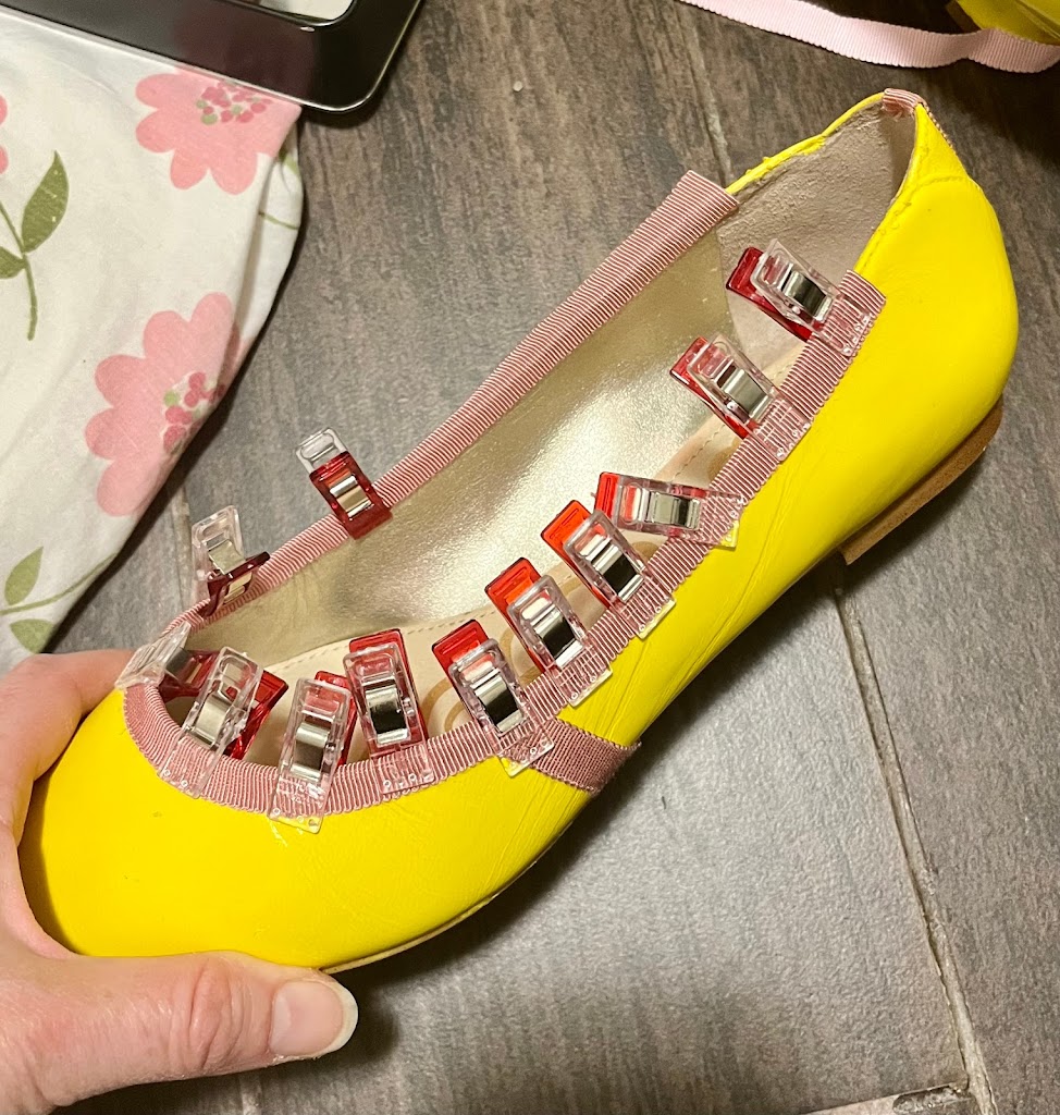 A bright yellow painted 1790s shoe with soft pink petersham ribbon being glued around the foot opening. The ribbon is held in place by a dozen red quilting clips
