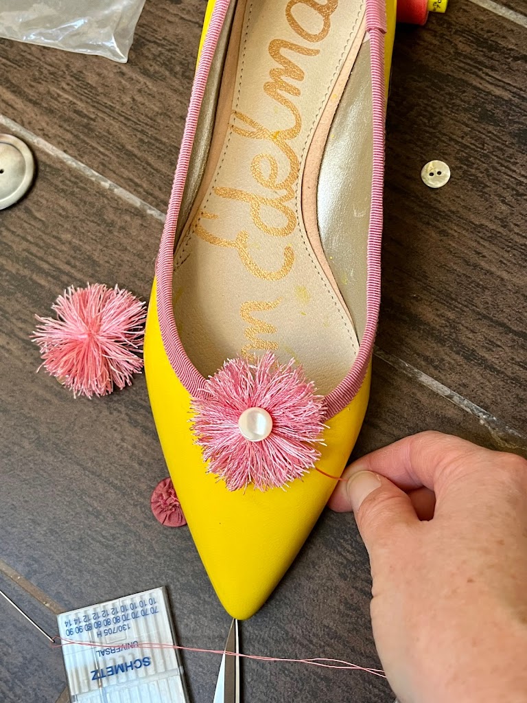 A close-up of a bright yellow 1790s shoe with a pink pom-pom on the toe. The pom-pom is centered with a white mother of pearl shank button