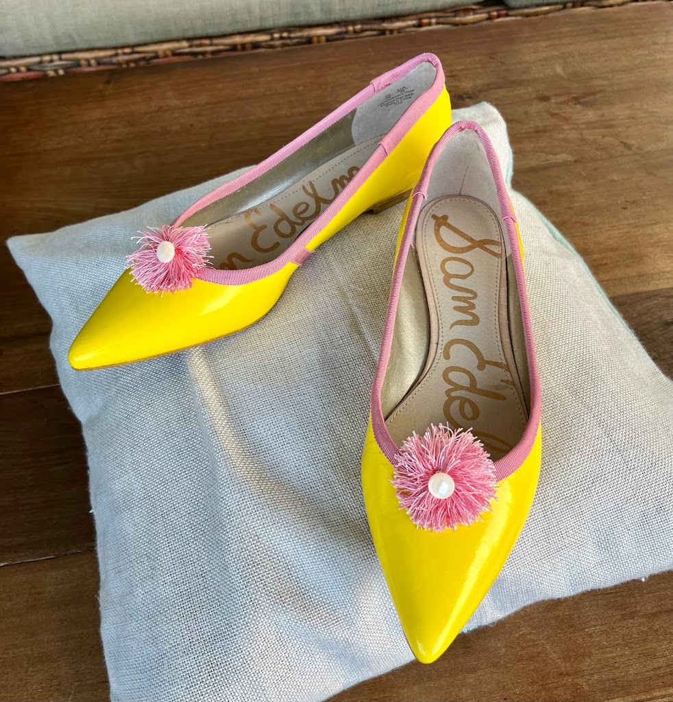 A pair of painted 1790s shoes sitting on a pale beige cushion. The shoes are a bright sunny, shiny yellow and bound in soft pink petersham ribbon. They have pink pompoms on the toes, each centered with a small white mother of pearl shank button.