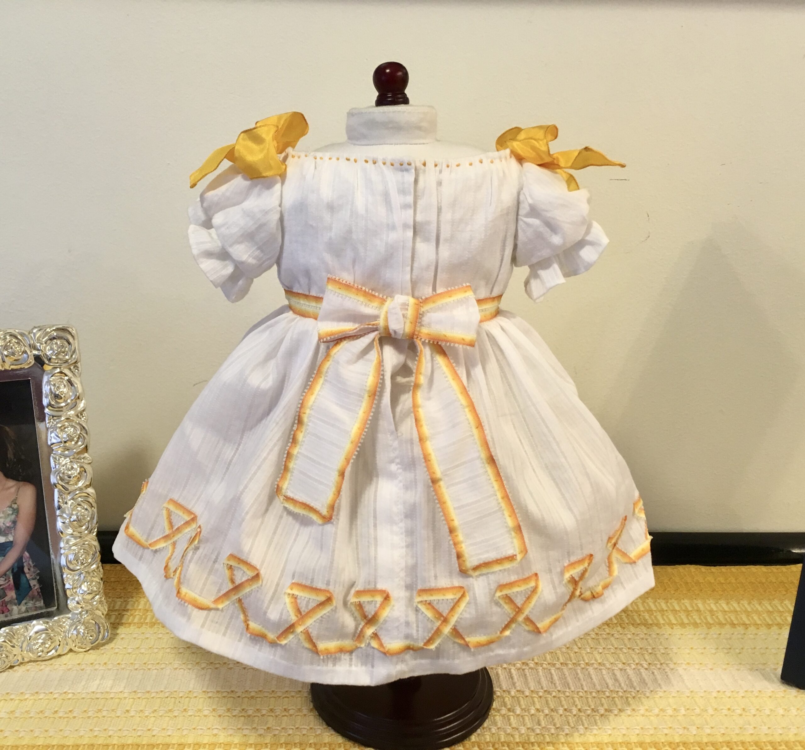 The front view of the Winterhalter princess dress - a white dress with a full skirt, gathered bodice front and puffed sleeves. There are yellow bows on the shoulders, yellow orange trimming on the skirt and a bow at the back waist.
