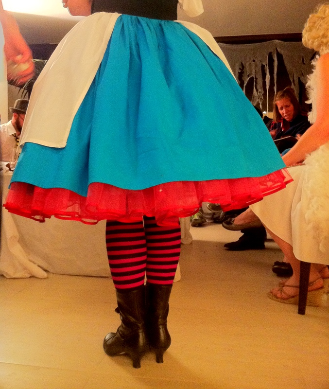 A view of a woman's back from the waist down. She is wearing a white apron and blue skirt over a pink net petticoat, pink striped stockings and black high-heeled boots.