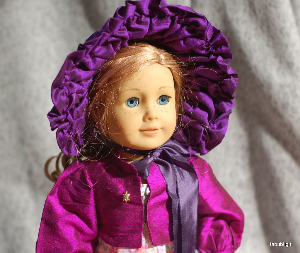 An american girl doll wears a purple silk spencer jacket and a historically inspired dark purple bonnet.