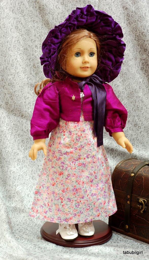 An American Girl Doll wears a regency ensemble of a floral dress, a purple silk spencer and a dark purple bonnet.
