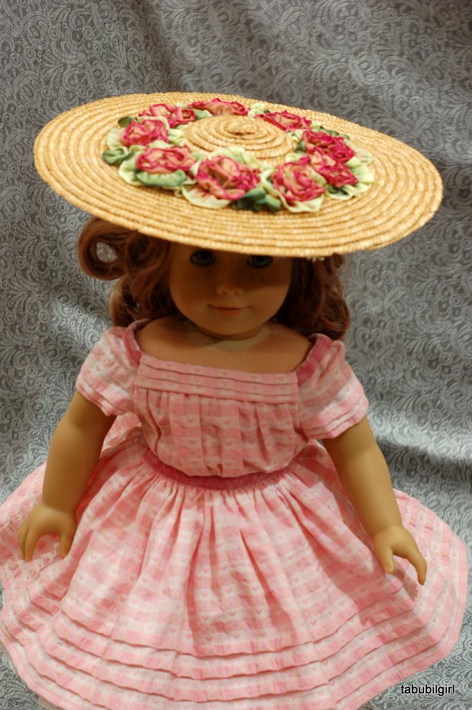 An American Girl doll wears a pink 1860s dress and a wide straw hat trimmed with roses