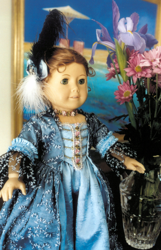 An American Girl Doll in an elaborate blue rococo gown stands in front of a painted backdrop