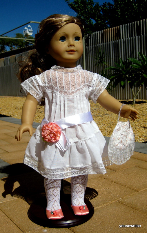 An American Girl Doll wears an Edwardian ensemble consisting of white dress, embroidered purse and white sash