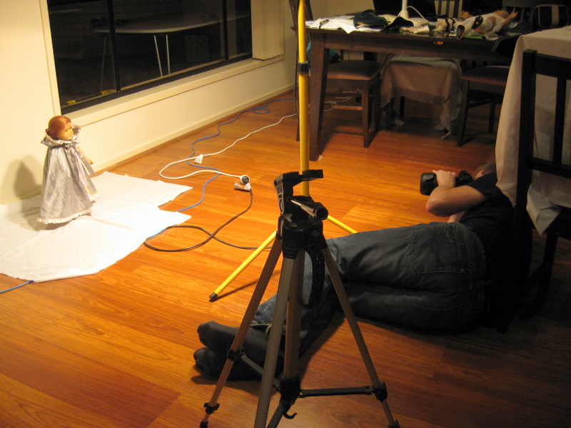 A man lies on the floor, holding a camera. in front of him a doll stands on a strip of fabric in a pool of light