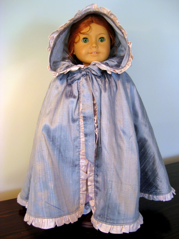 An American Girl doll wears a long silver cape with a pink ruffle.