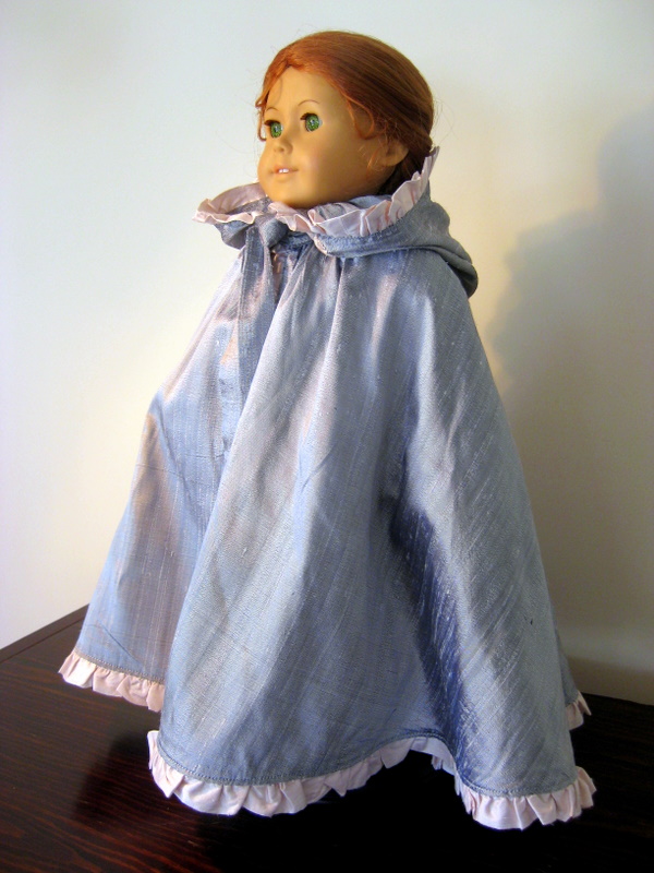A 3/4 view of an American Girl doll wearing a long silver cloak with a pink ruffle.