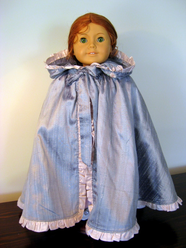 An American Girl doll wears a long silver cloak with a pink ruffle. The hood is folded back away from her face.