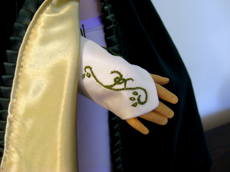 A doll arm wearing an embroidered silk mitt peeks out from the folds of a green and gold cloak.