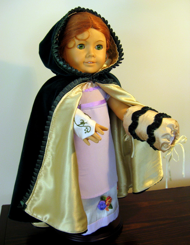 An American Girl Doll wearings a Green cloak, white mitts and gold muff over a purple silk gown
