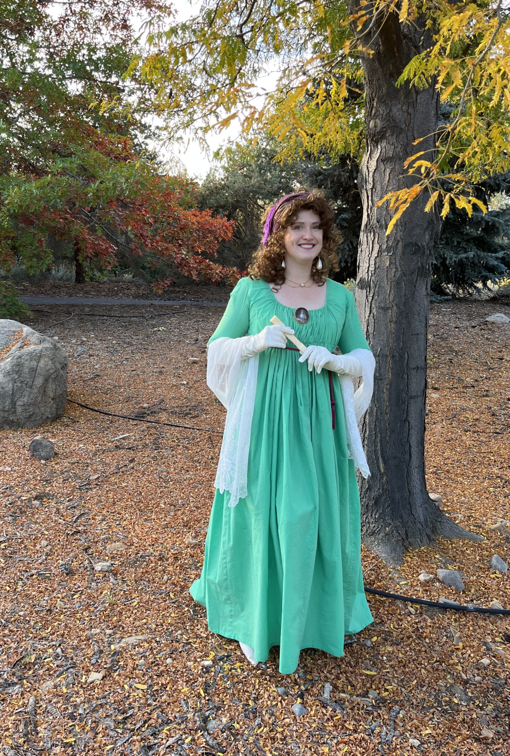 Tabubilgirl in a green 1790s round gown stand beneath a tree holding a vintage celluloid fan.