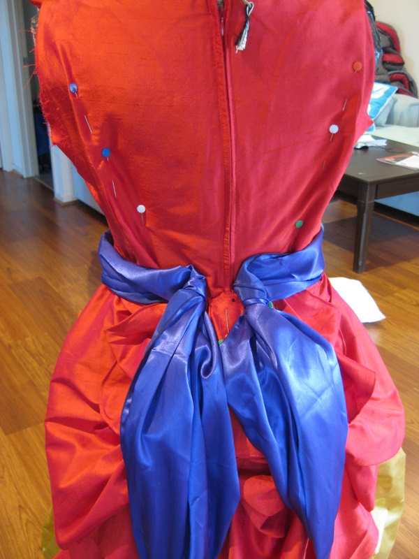 detail of a purple sash pinned to the waist of a red silk ballgown