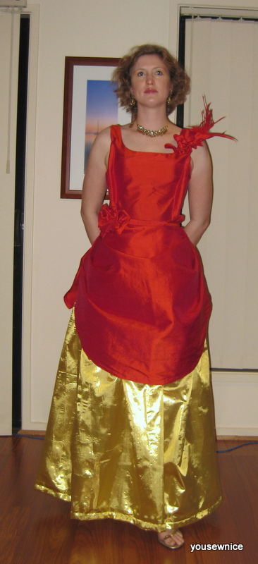 Tabubilgirl stands in a hallway. She is wearing a ballgown consisting of a red silk gown draped over a gold lame petticoat.