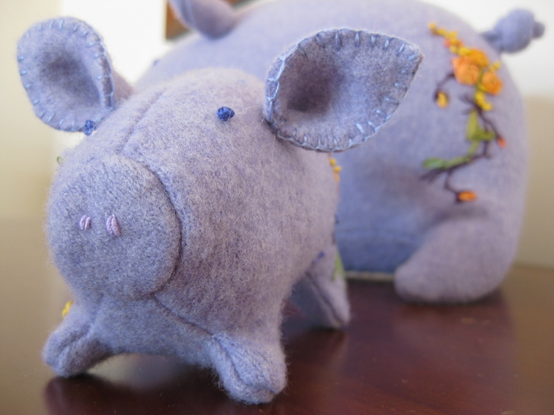 a purple stuffed pig faces the camera. In the background, a second pig shows his embroidered bottom.