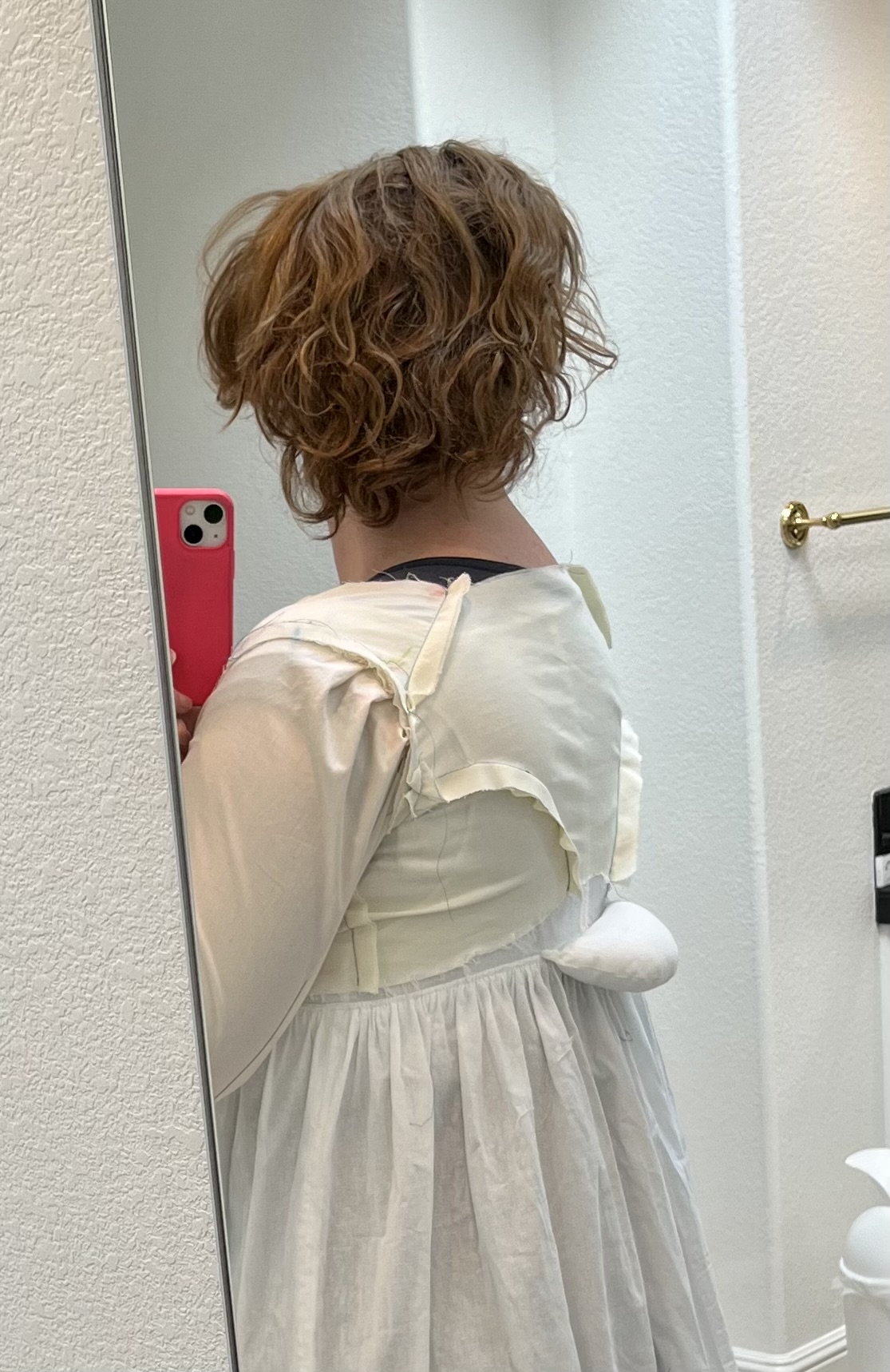 A woman faces away from the camera. She is wearing a white petticoat with a small bum roll and a white 1790s bodice mockup