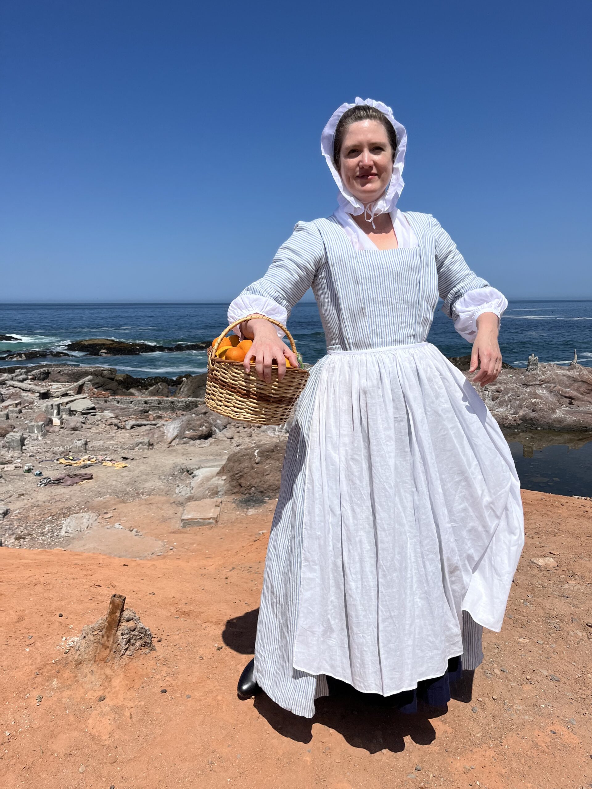Tabubilgirl stands on a rocky seashore. She wears a 1750s striped linen english nightgown and a white linen lappet cap and carries a basket of oranges.