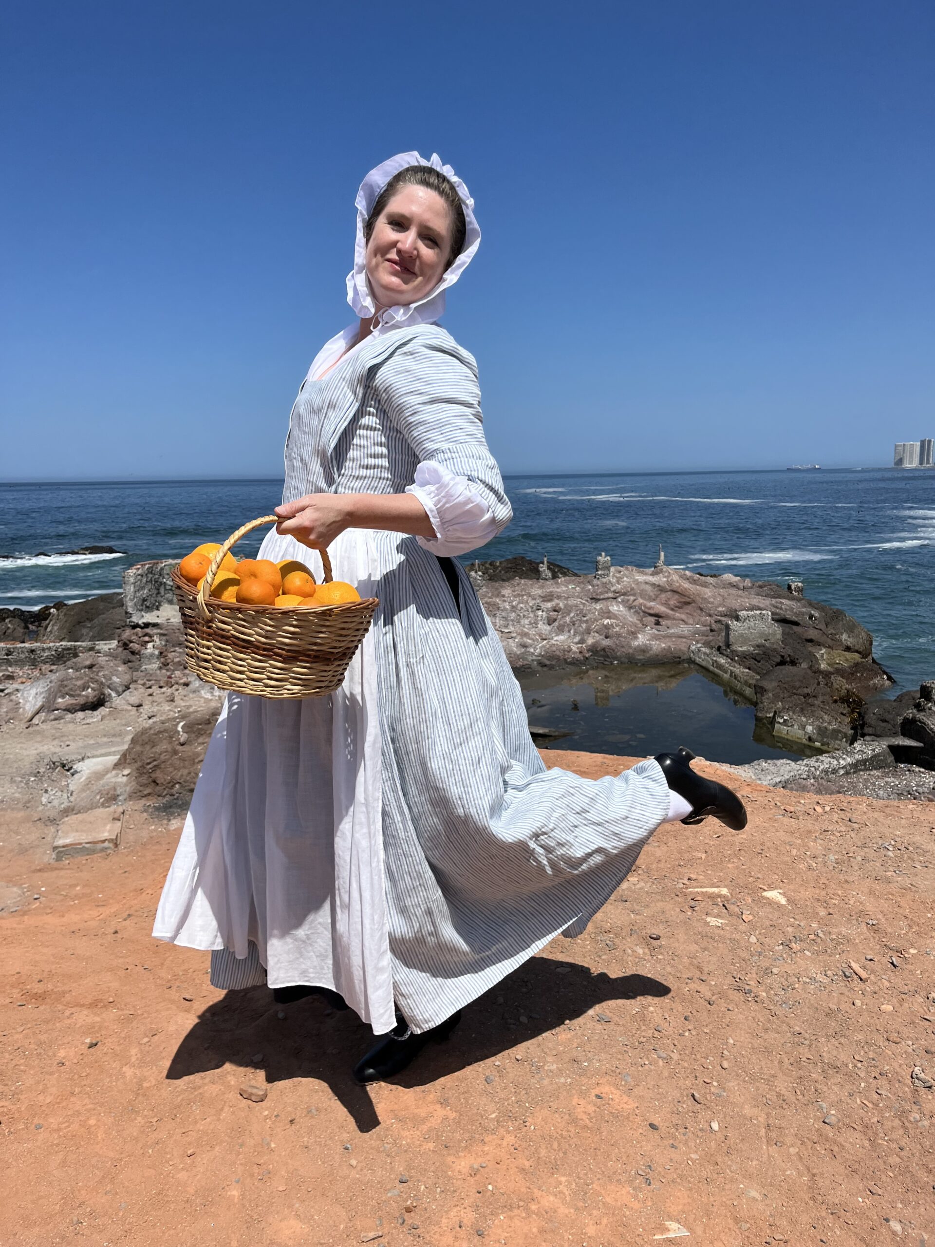 Tabubilgirl dances on a barren, rocky seashore. She wears a striped linen English nightgown and a white linen lappet cap and carries a basket of oranges.