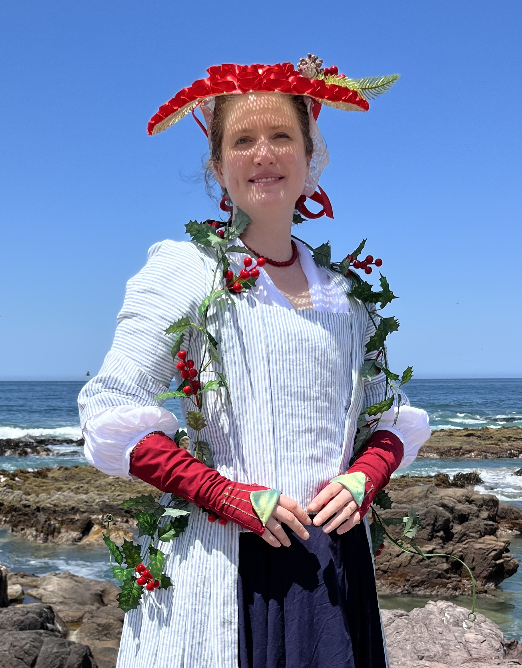 Tabubilgirl stands on a rocky shore. She is dressed for a tropical christmas and is wearing a striped linen English nightgown and a blue cotton petticoat. She is holding an 18th Century Bergere Hat . The hat sits high and flat on her head.