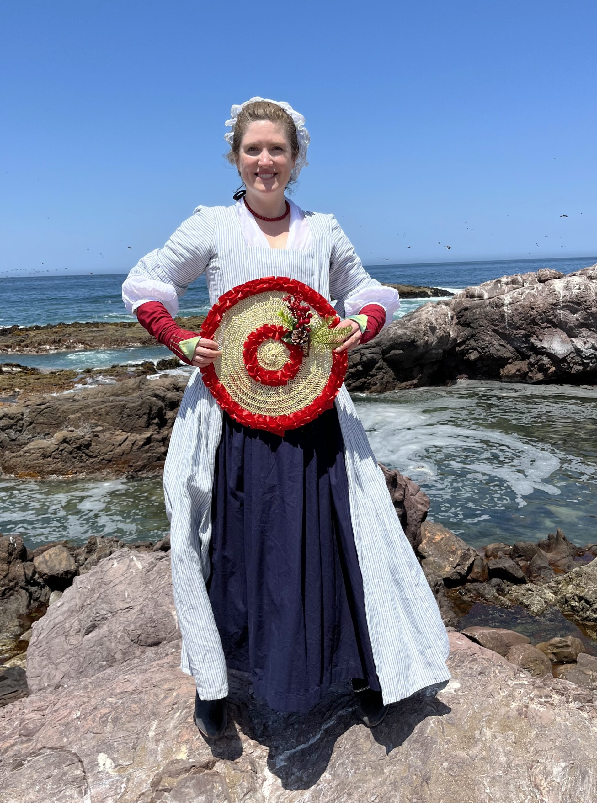 Tabubilgirl stands on a rocky shore. She is wearing a striped linen english nightgown and a blue cotton petticoat. She is holding an 18th Century Bergere Hat decorated for Christmas