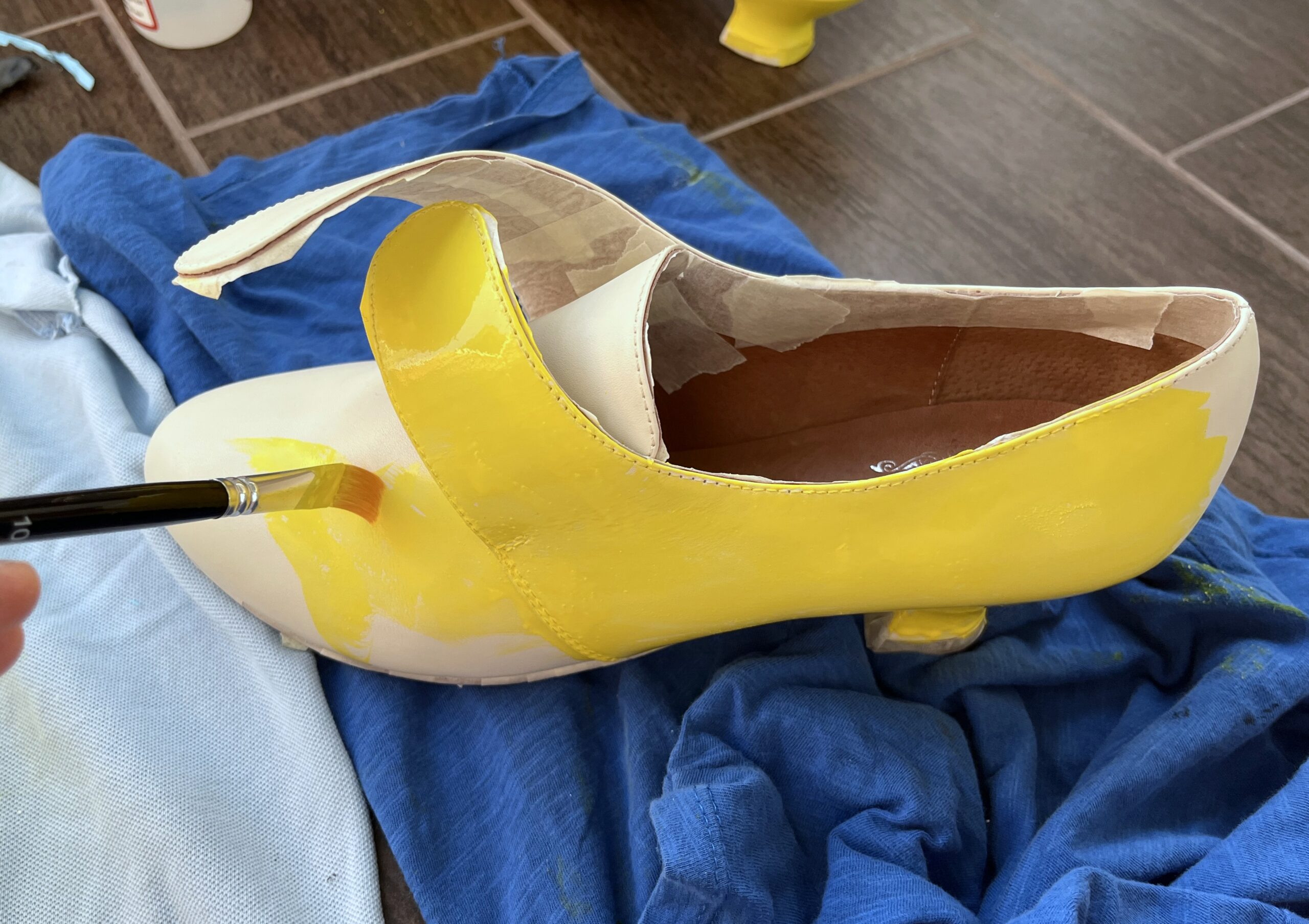 A brush painting streaks of bright yellow paint onto the toe of a white American Duchess Kensington shoe.