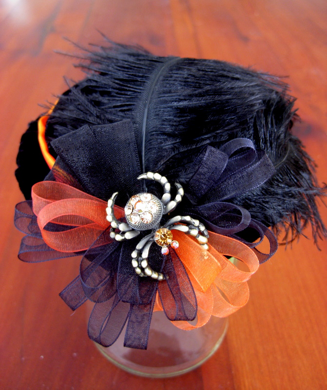 A black American Girl doll hat is decorated with black feathers, orange ribbons and a rhinestone spider