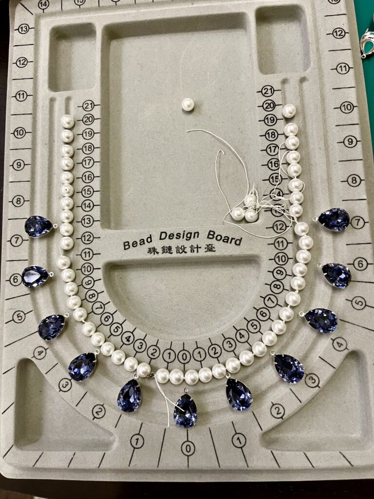 An in-progress pearl necklace lies on a beading board, with purple crystal drops lying in place next to the pearls.