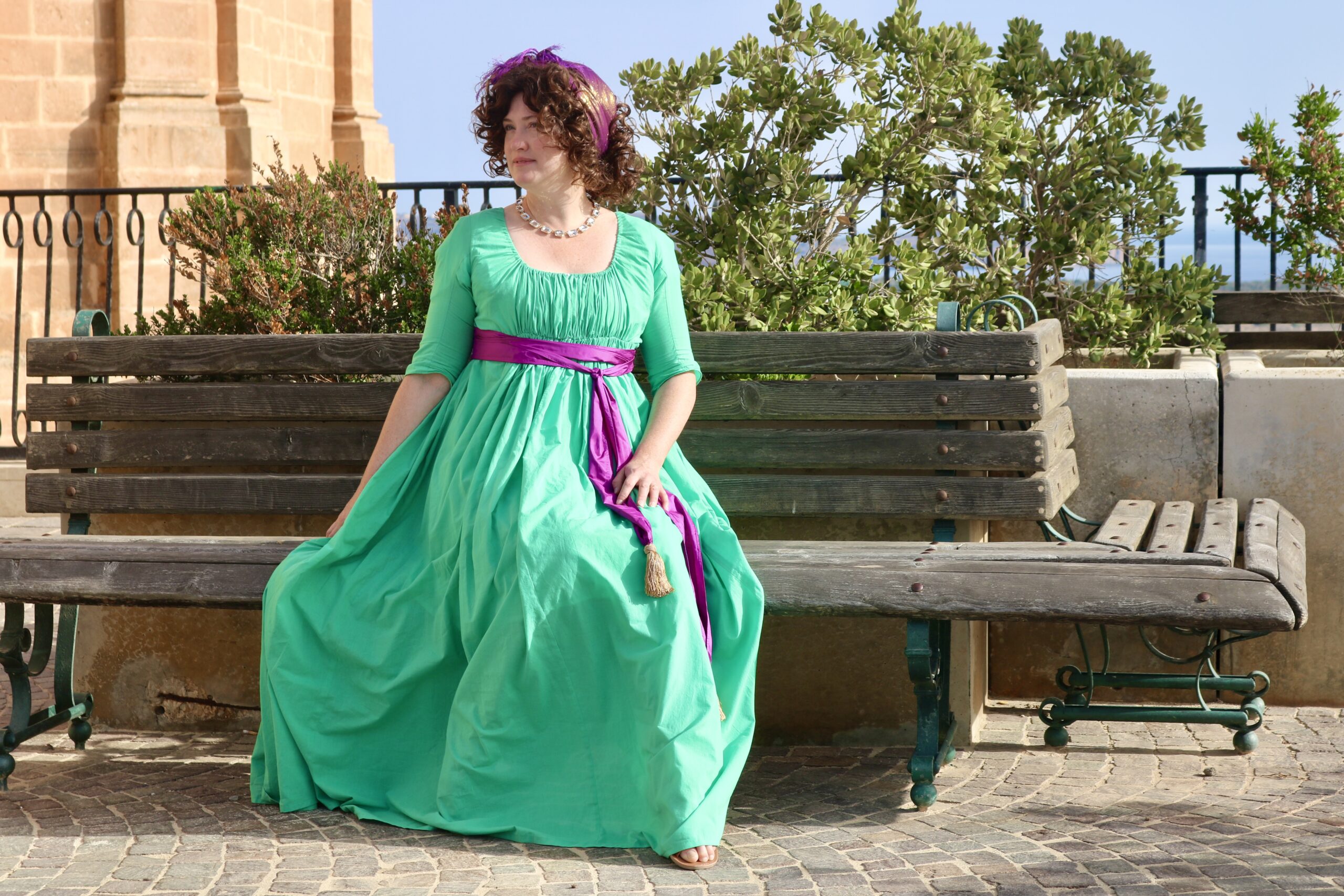 Tabubilgirl sits on a bench. She is wearing a green 1790s round gown with a purple sash.