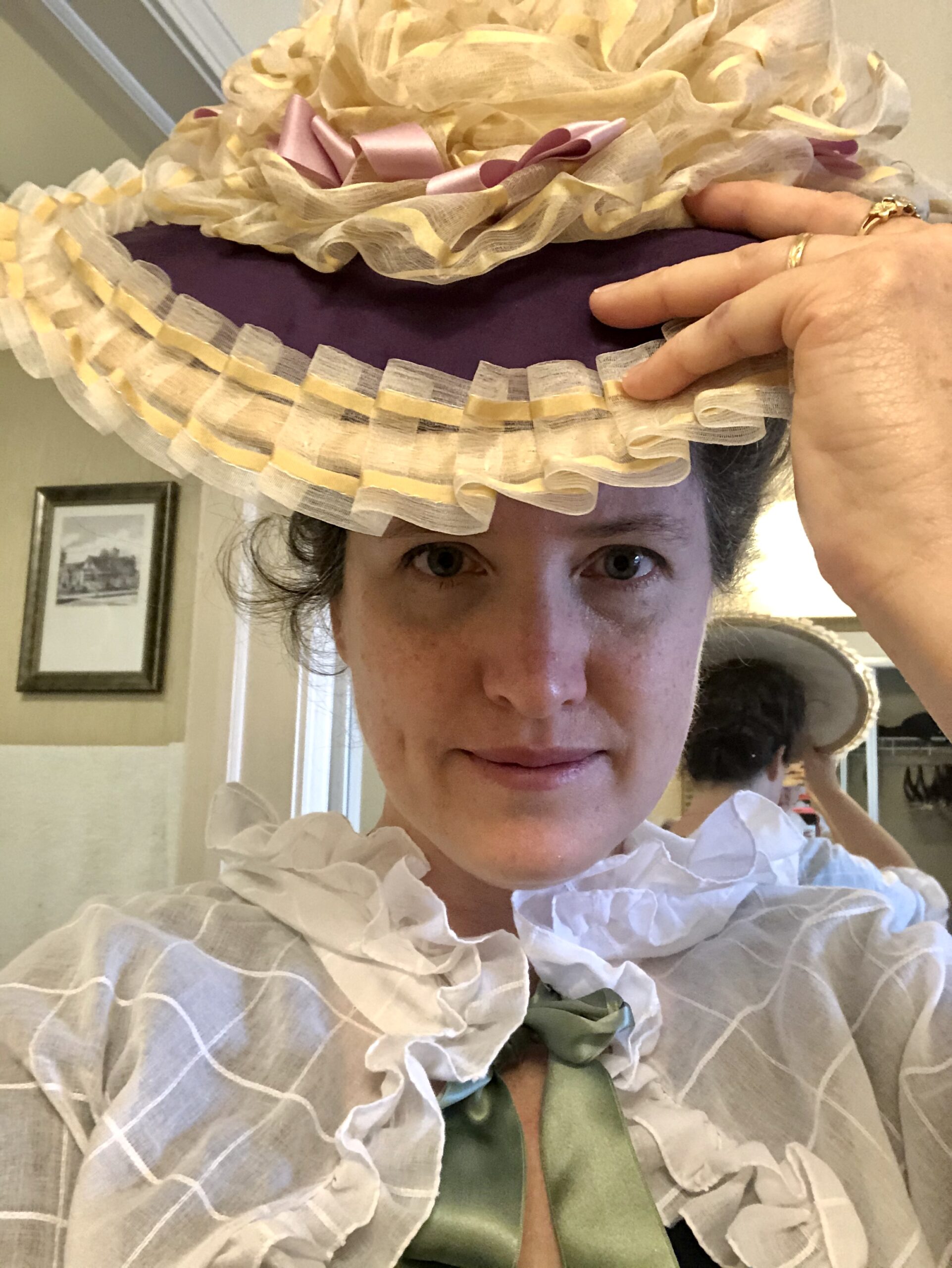 A woman smiles at the camera. She is lifting up a purple bergere hat trimmed with gold silk gauze and pink ribbons.