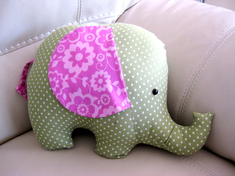 a green stuffed elephant with pink ears sits on a sofa.