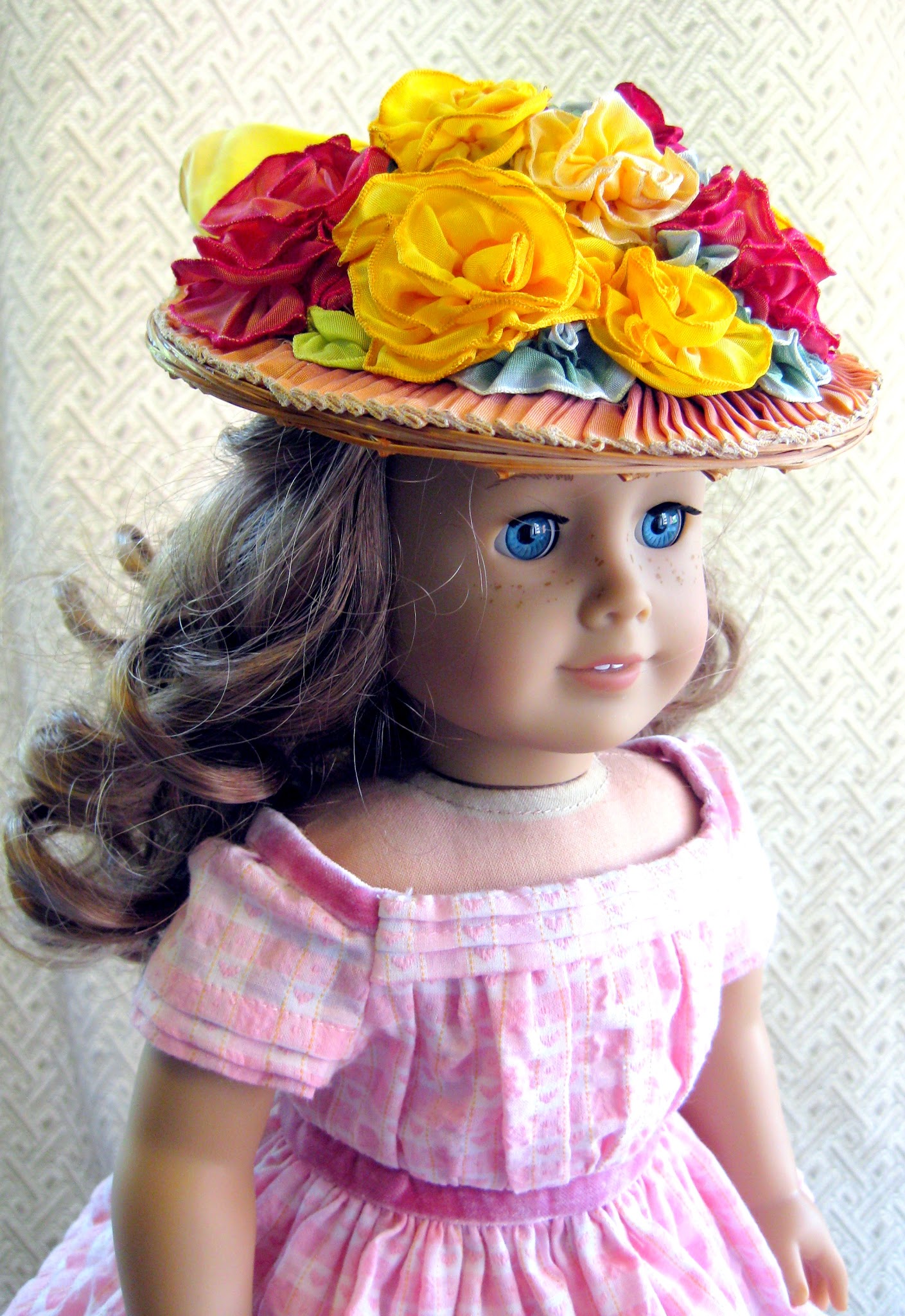 An American Girl doll in a pink dress wears a straw hat covered in wire-ribbon roses