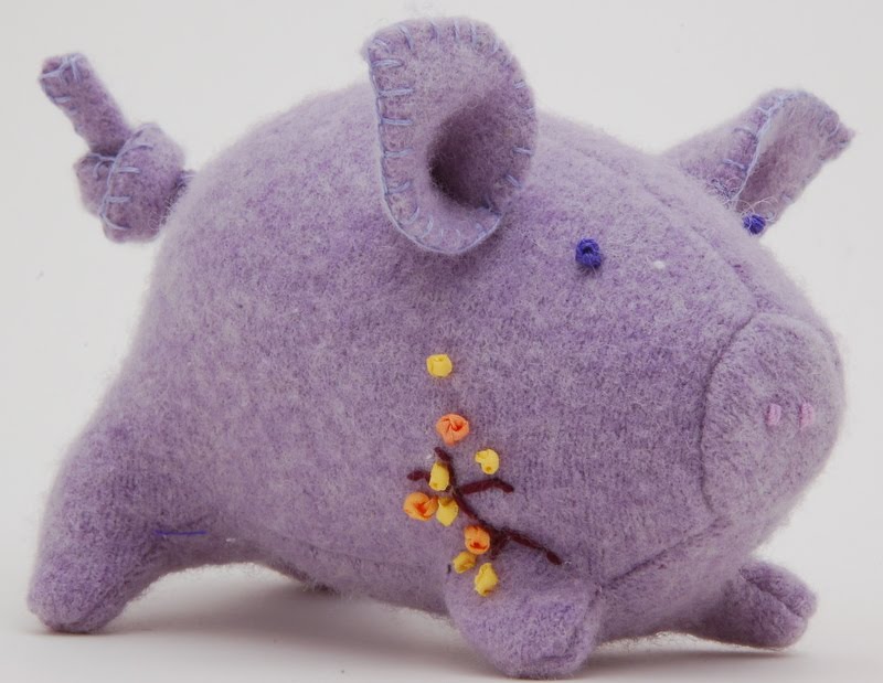 A stuffed wool pig with embroidery on his side faces the camera