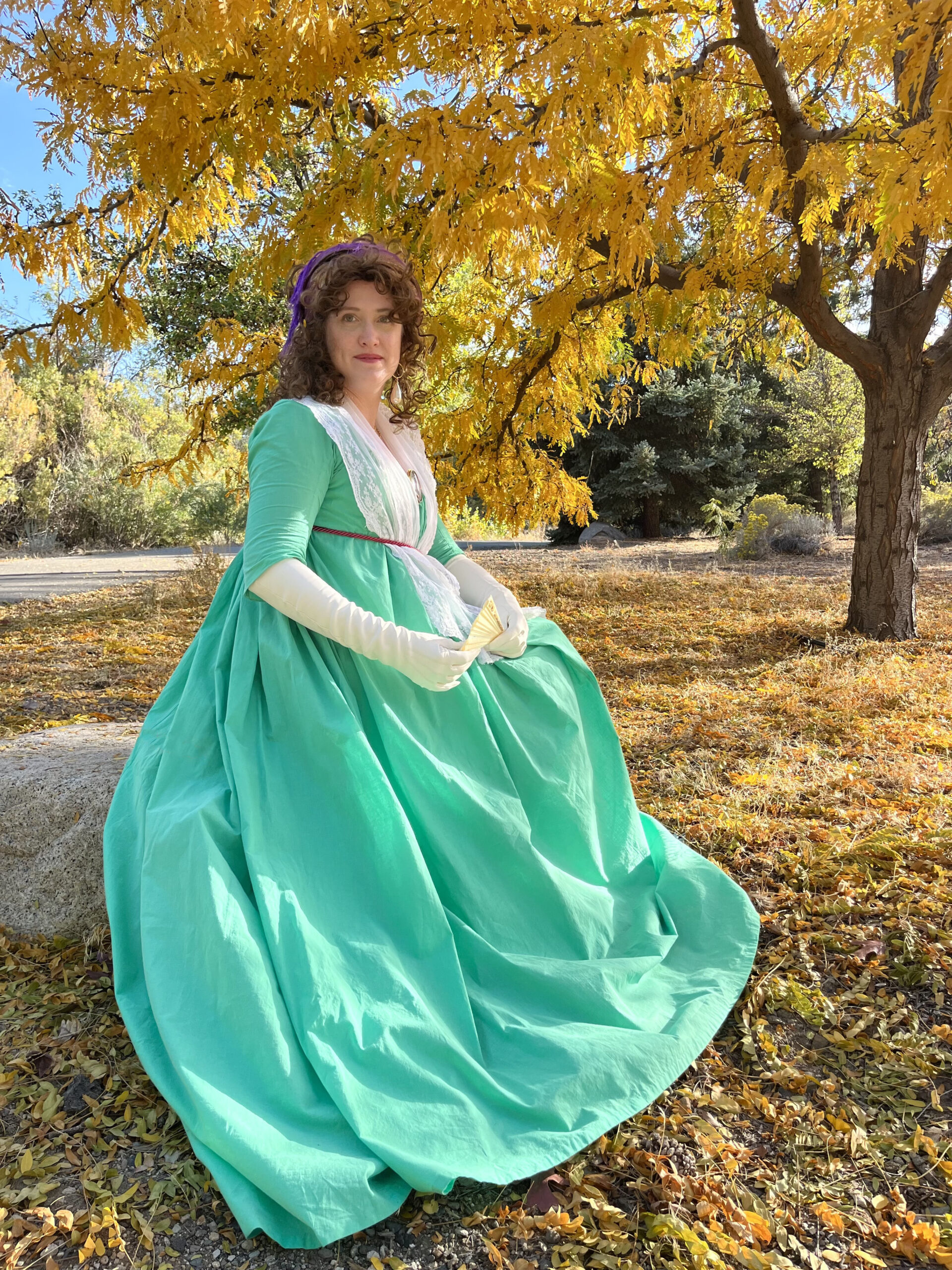 Tabubilgirl in a green 1790s round gown sits beneath a tree with gold autumn leaves.