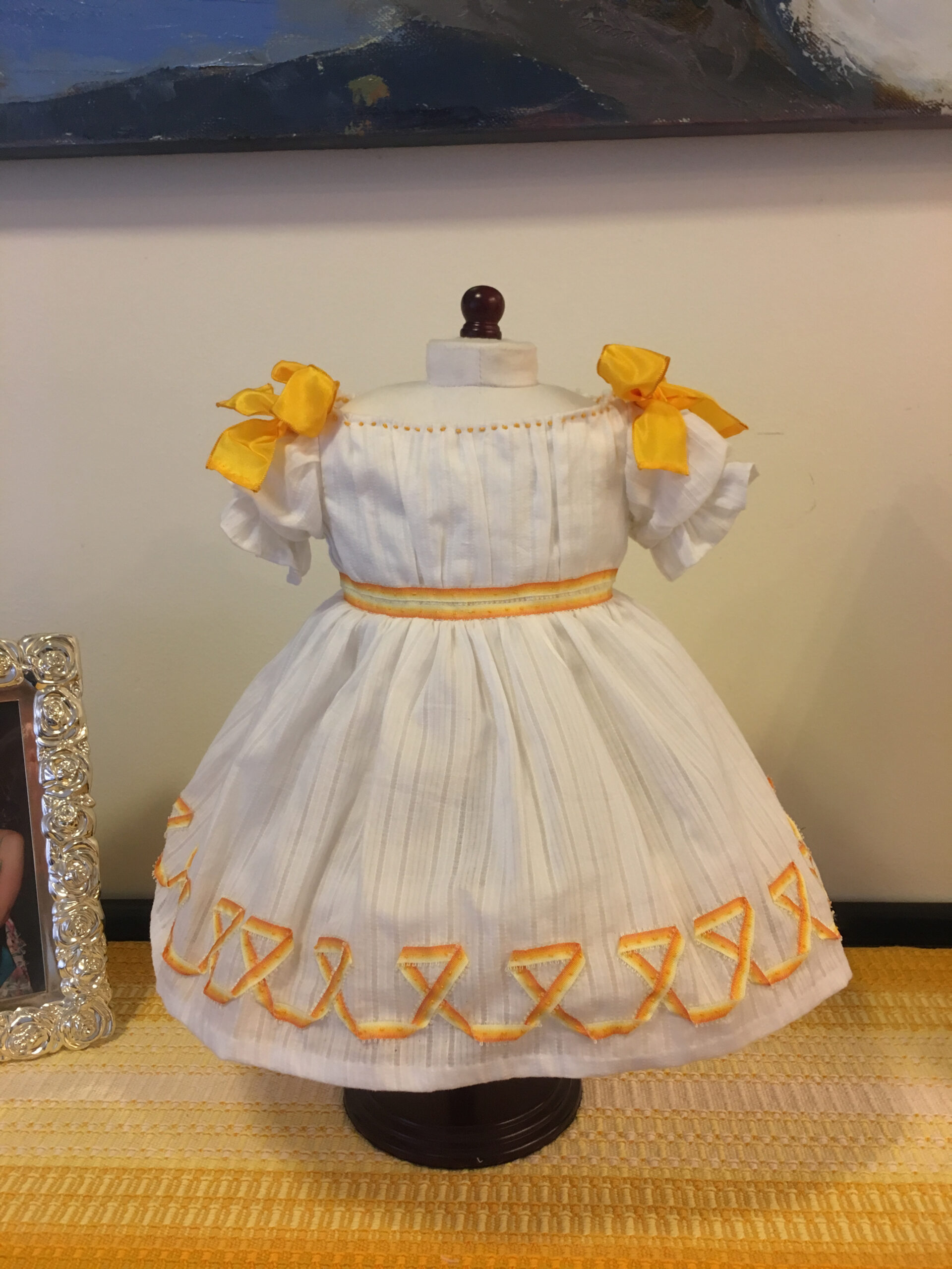 The front view of the Winterhalter princess dress - a white dress with a full skirt, gathered bodice front and puffed sleeves. There are yellow bows on the shoulders and yellow orange trimming on the skirt and waist.