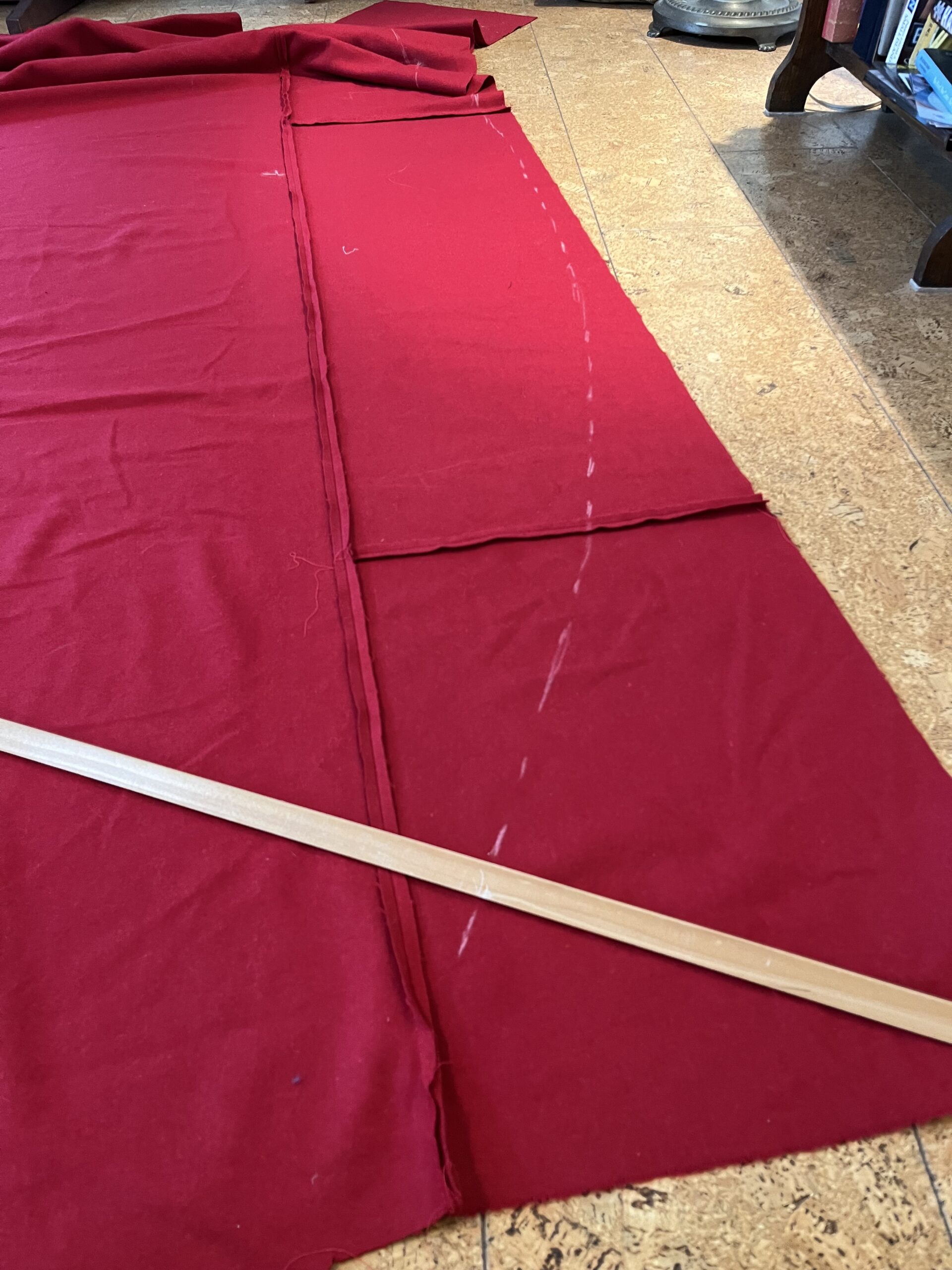 A long wooden rod is being used to trace a large curve across a pieced length of red wool fabric