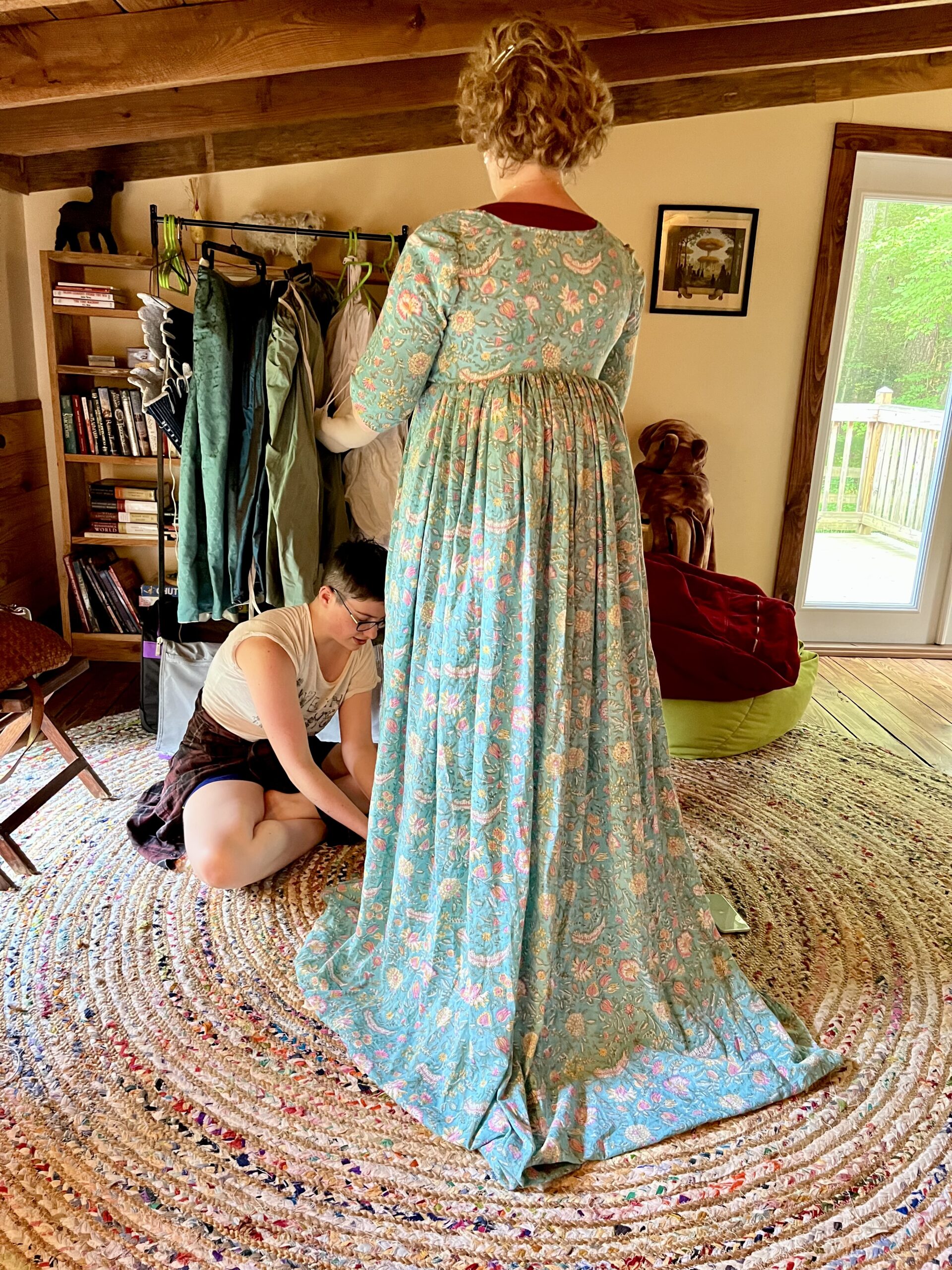 A woman is sitting on  a floor marking the hem of a green 1790s gown. The gown is held out by an extremely large bustle.