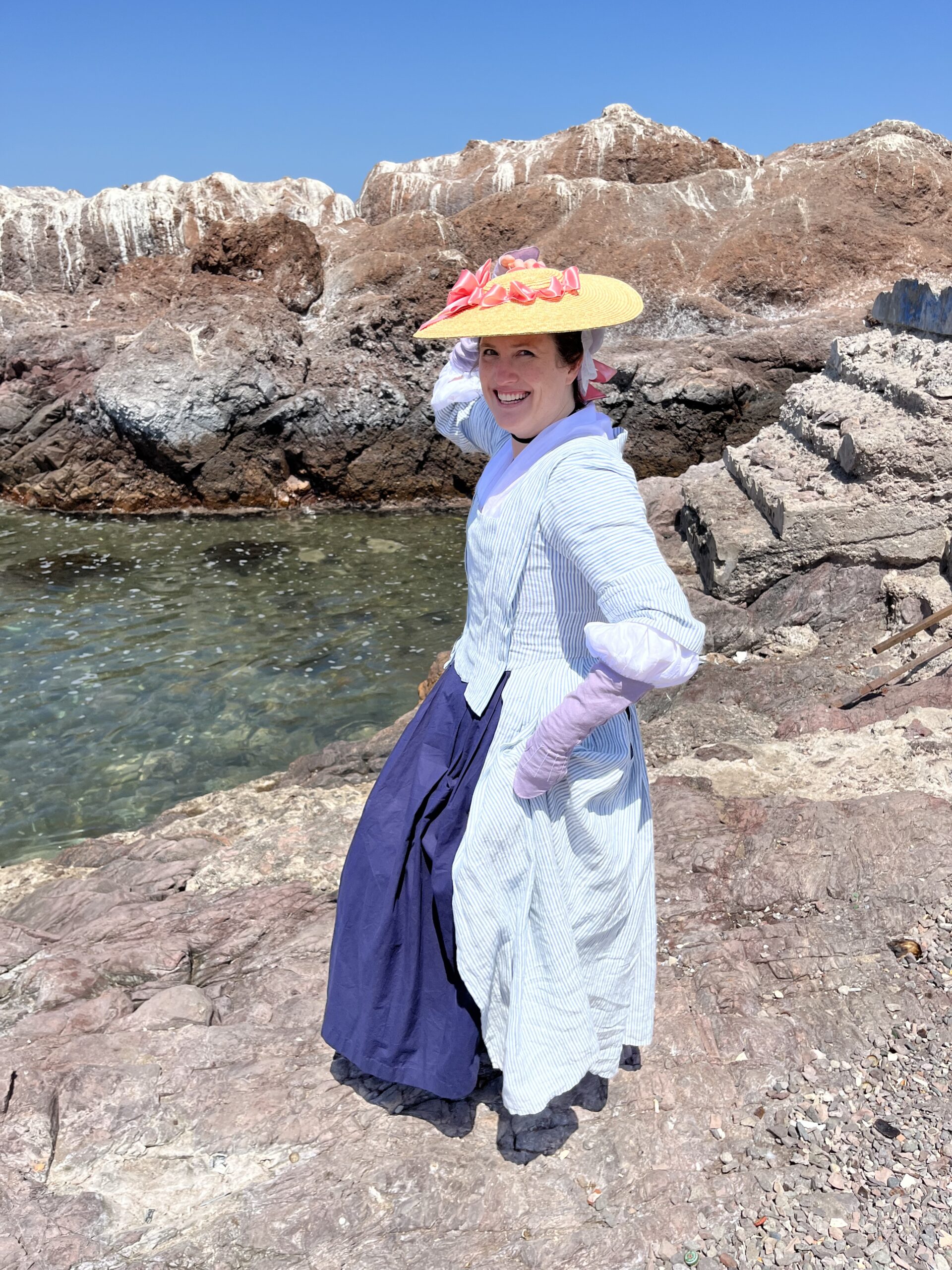 Tabubilgirl stands on a shore in the Atacama Desert, holding her hat onto her head. she wears a blue striped 1750s english nightgown, purple linen mitts and a straw hat trimmed with a pink ribbon