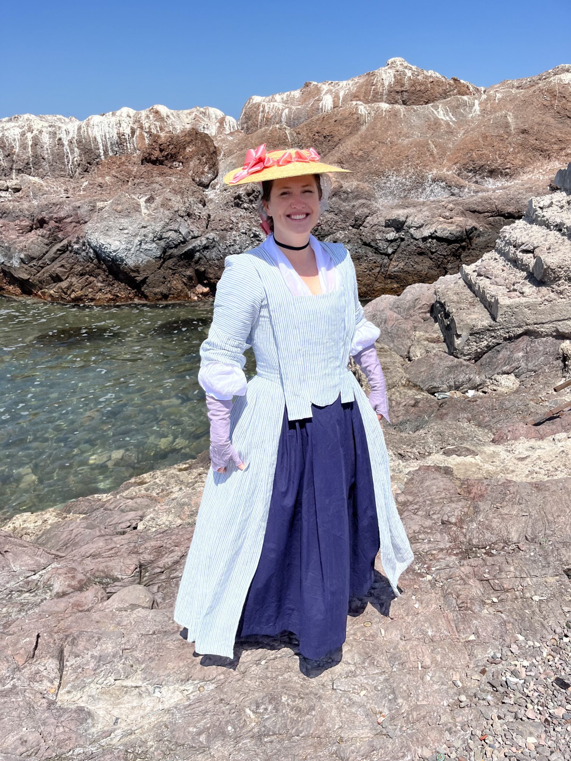 Tabubilgirl stands on a shore in the Atacama Desert. she wears a blue striped 1750s english nightgown, purple linen mitts and a straw hat trimmed with a pink ribbon
