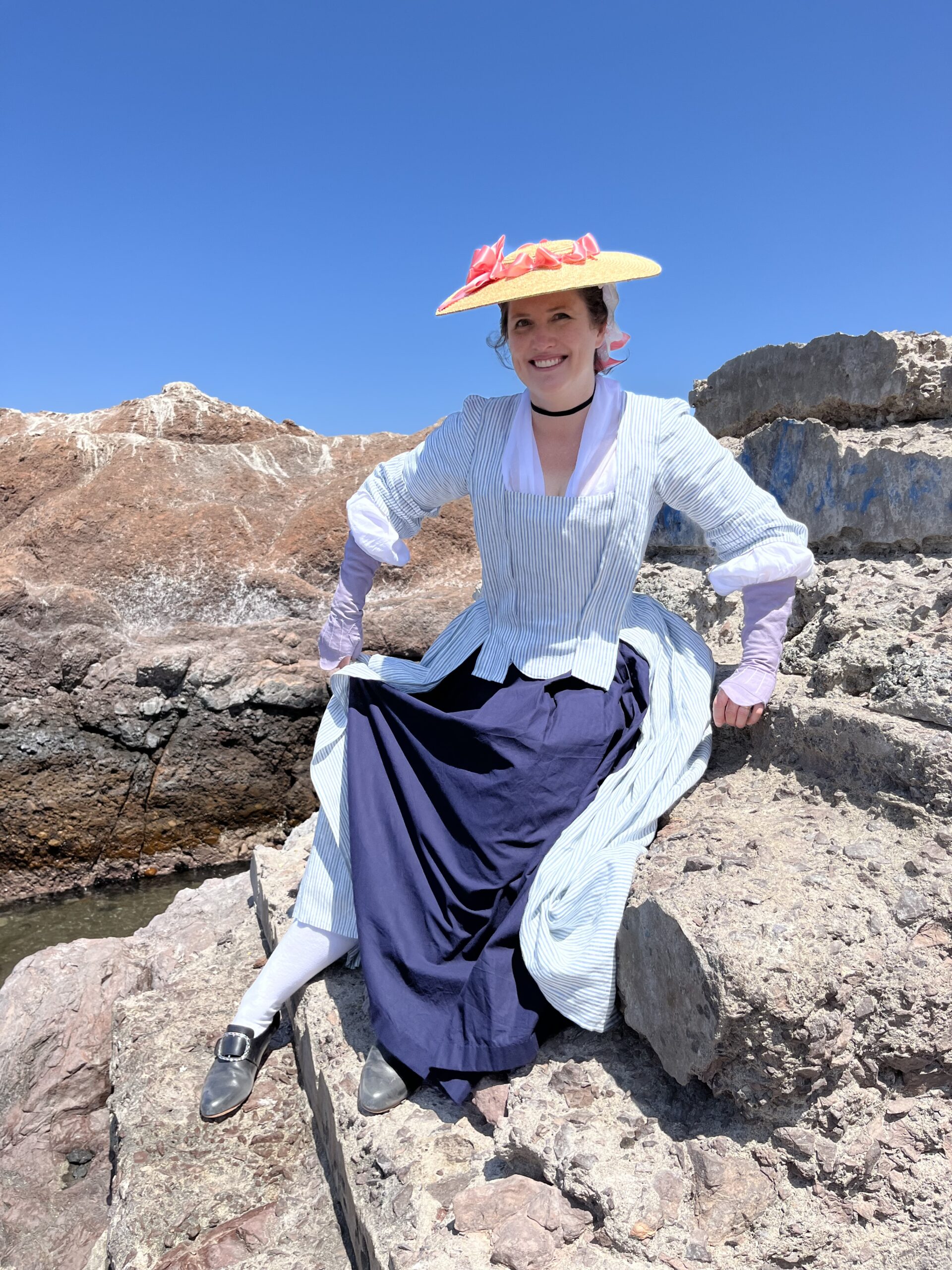 Tabubilgirl is seated on a rock in Northern Chile. she is wearing a striped 1750s New Year ensemble - an english nightgown, a blue cotton petticoat and a straw bergere hat trimmed with pink ribbons