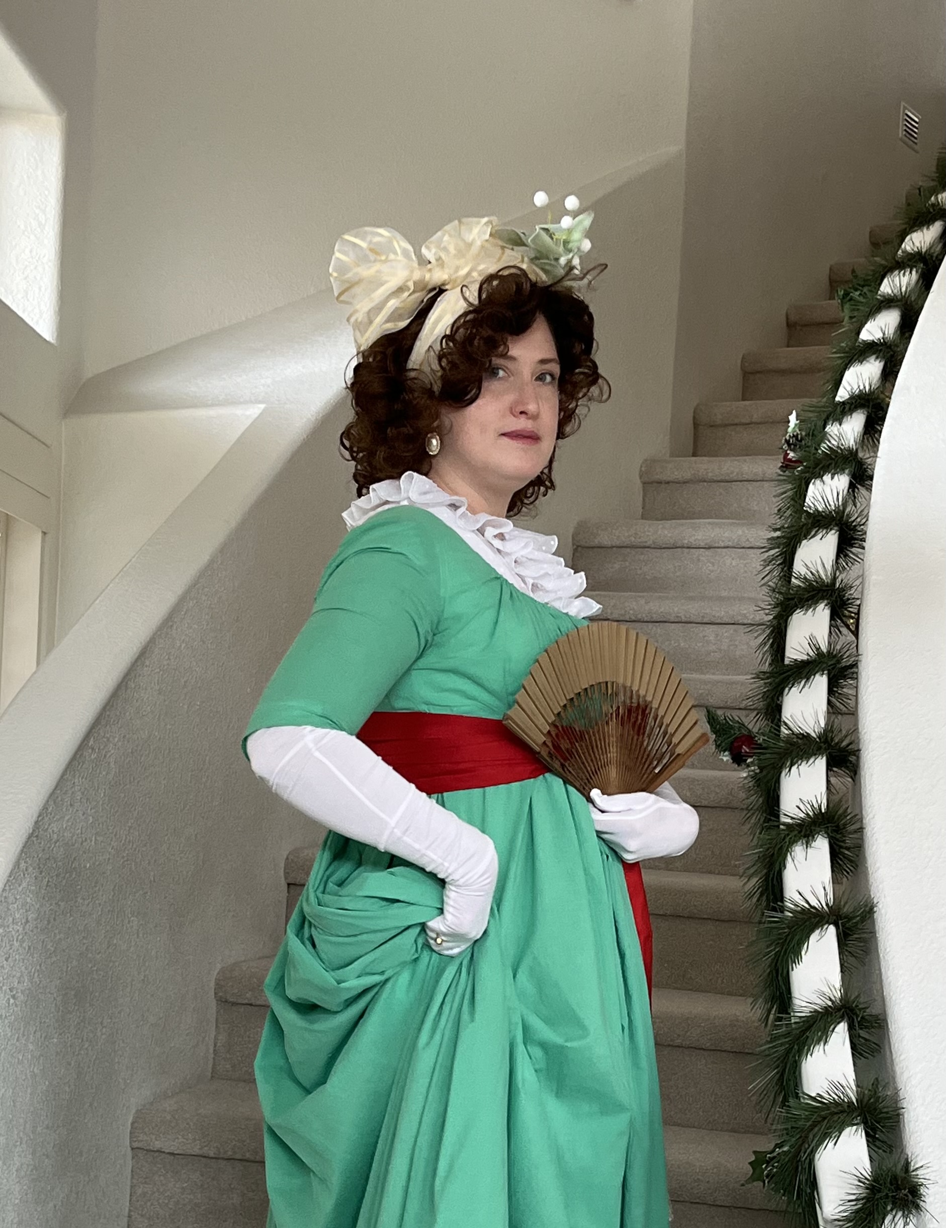 Tabubilgirl stands on a staircase wearing a 1790s New Year ensemble - a green 1790s round gown with a red sash and holding a gold fan.