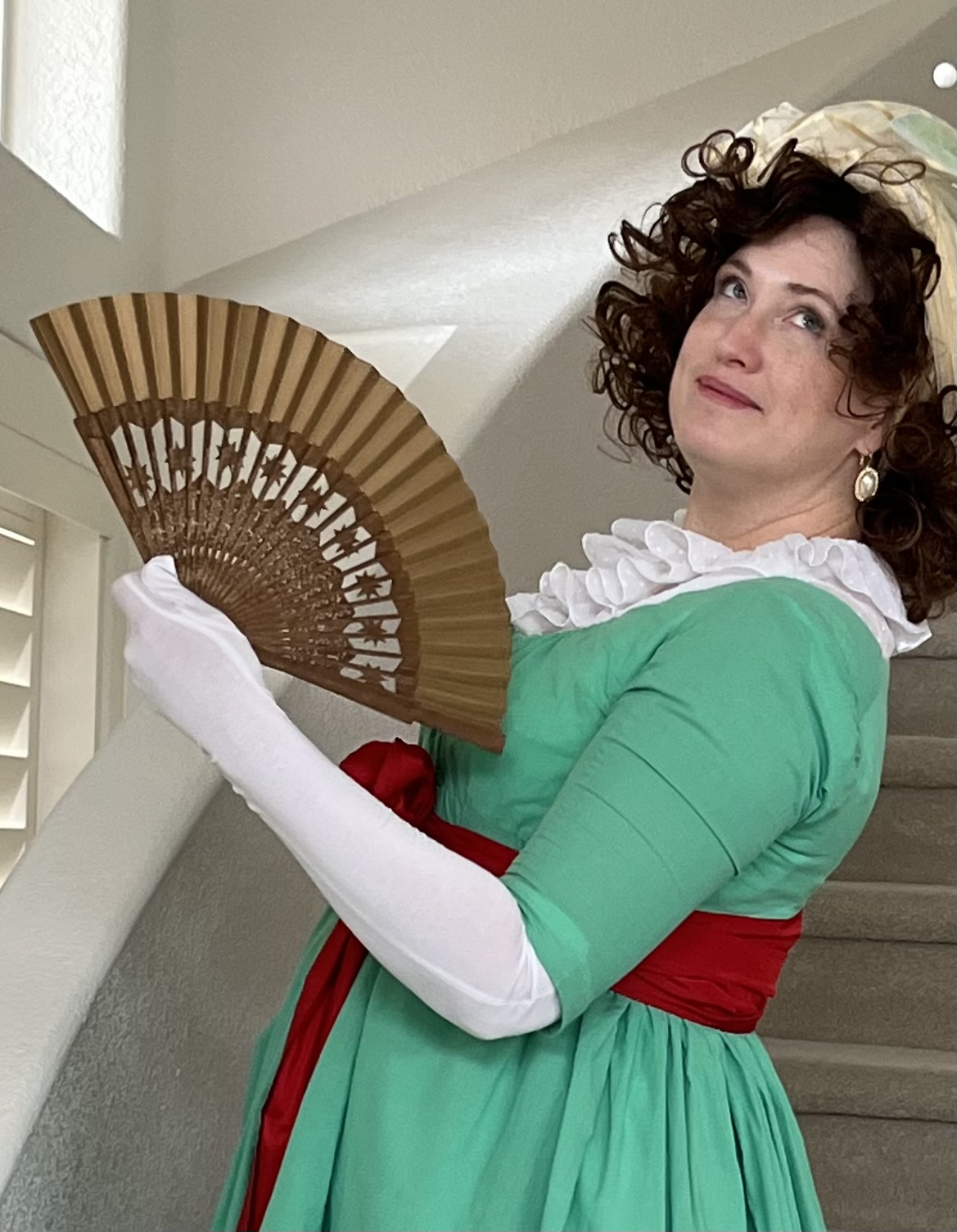 Tabubilgirl stands on a staircase wearing a green New Year gown with a white chemisette and a red sash and holding a gold fan.