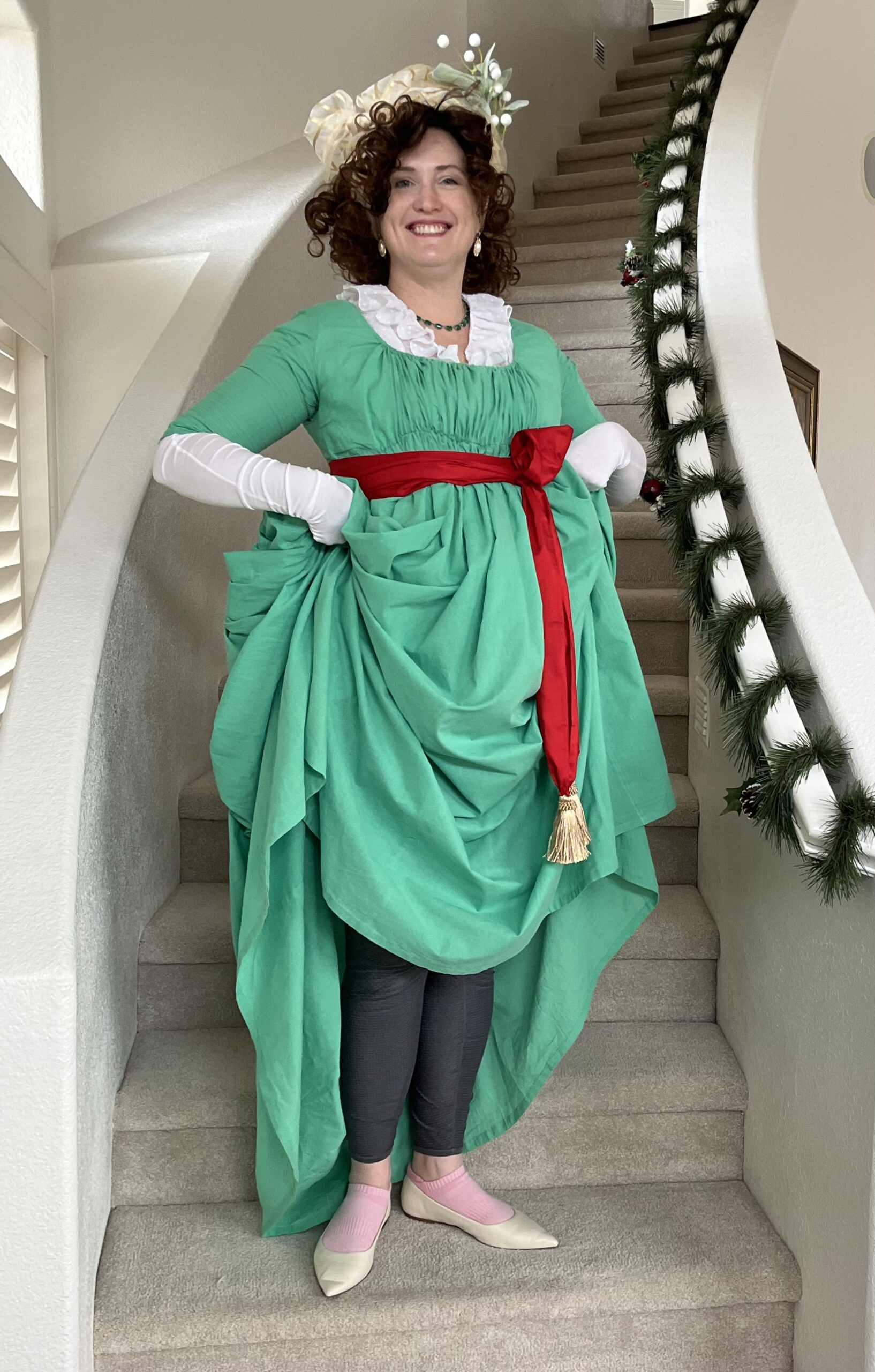 Tabubilgirl stands on a flight of stairs. She wears a green 1790s round gown. she is lifting up the skirts to show off modern thermal underwear