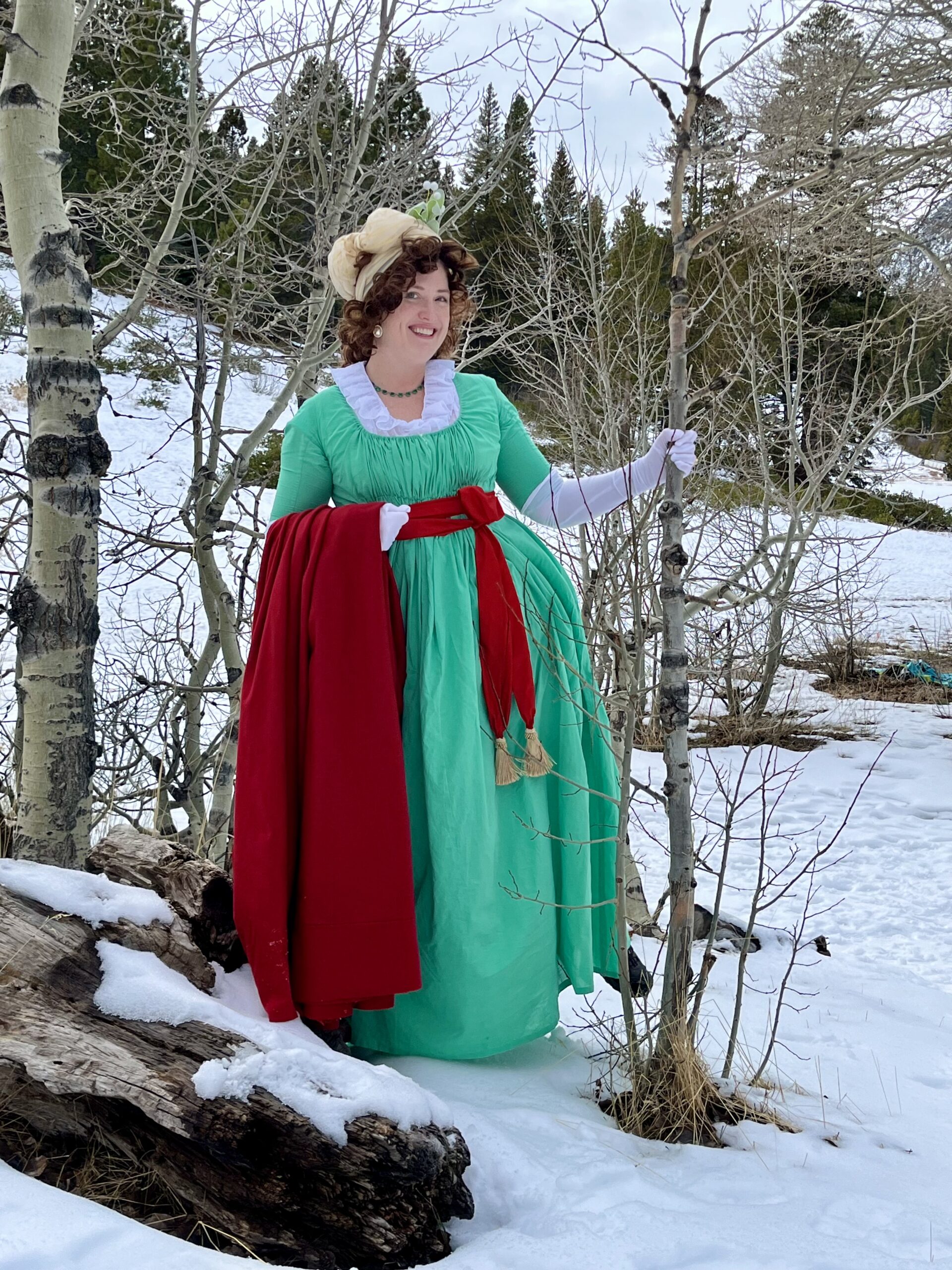 Tabubilgirl stands in a snowfield wearing a green 1790s round gown and a white chemisette and gloves. She carries a red cardinal cloak over her arm