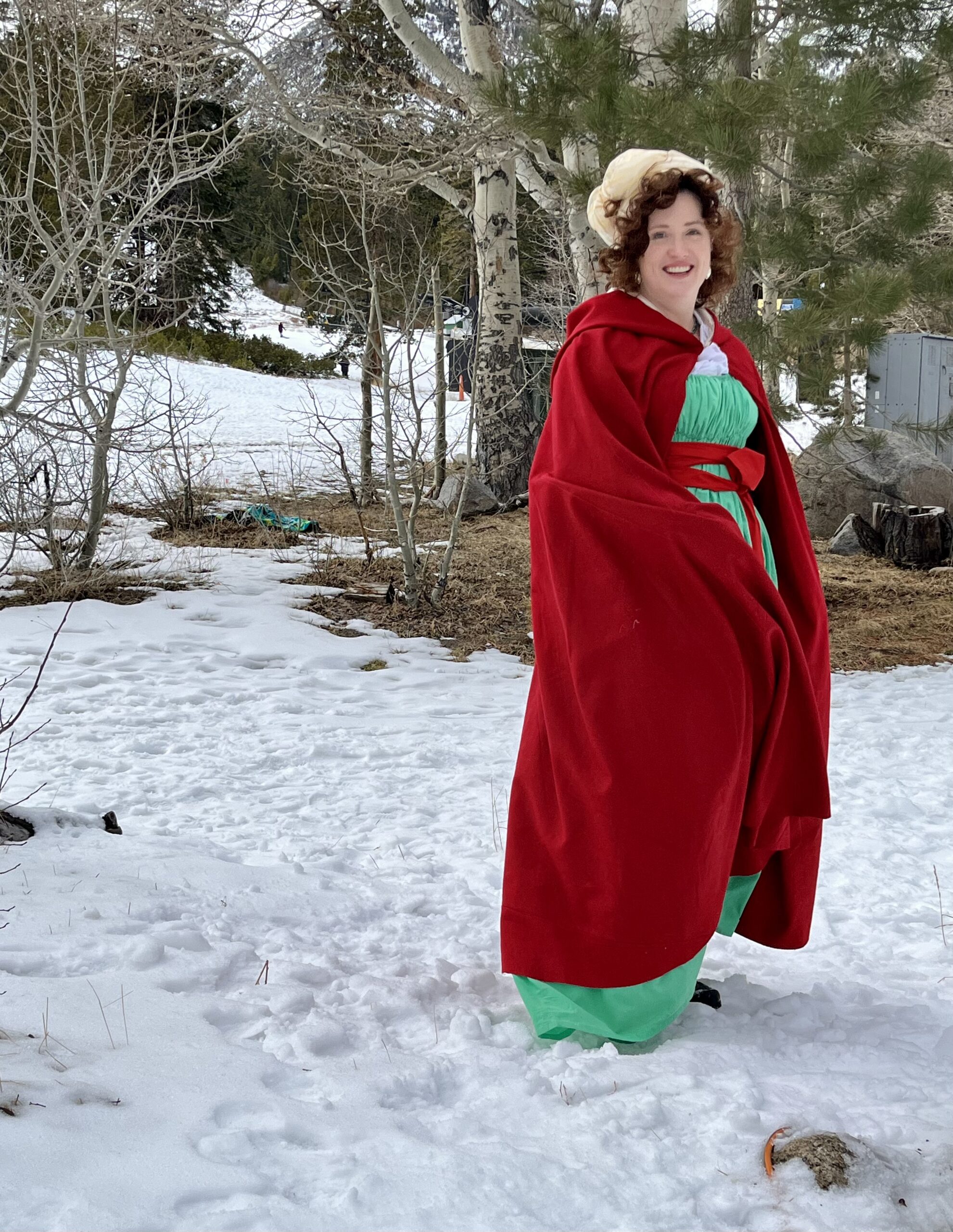 Tabubilgirl walks toward the camera wearing a green 1790s round gown and wrapped in a red cardinal cloak