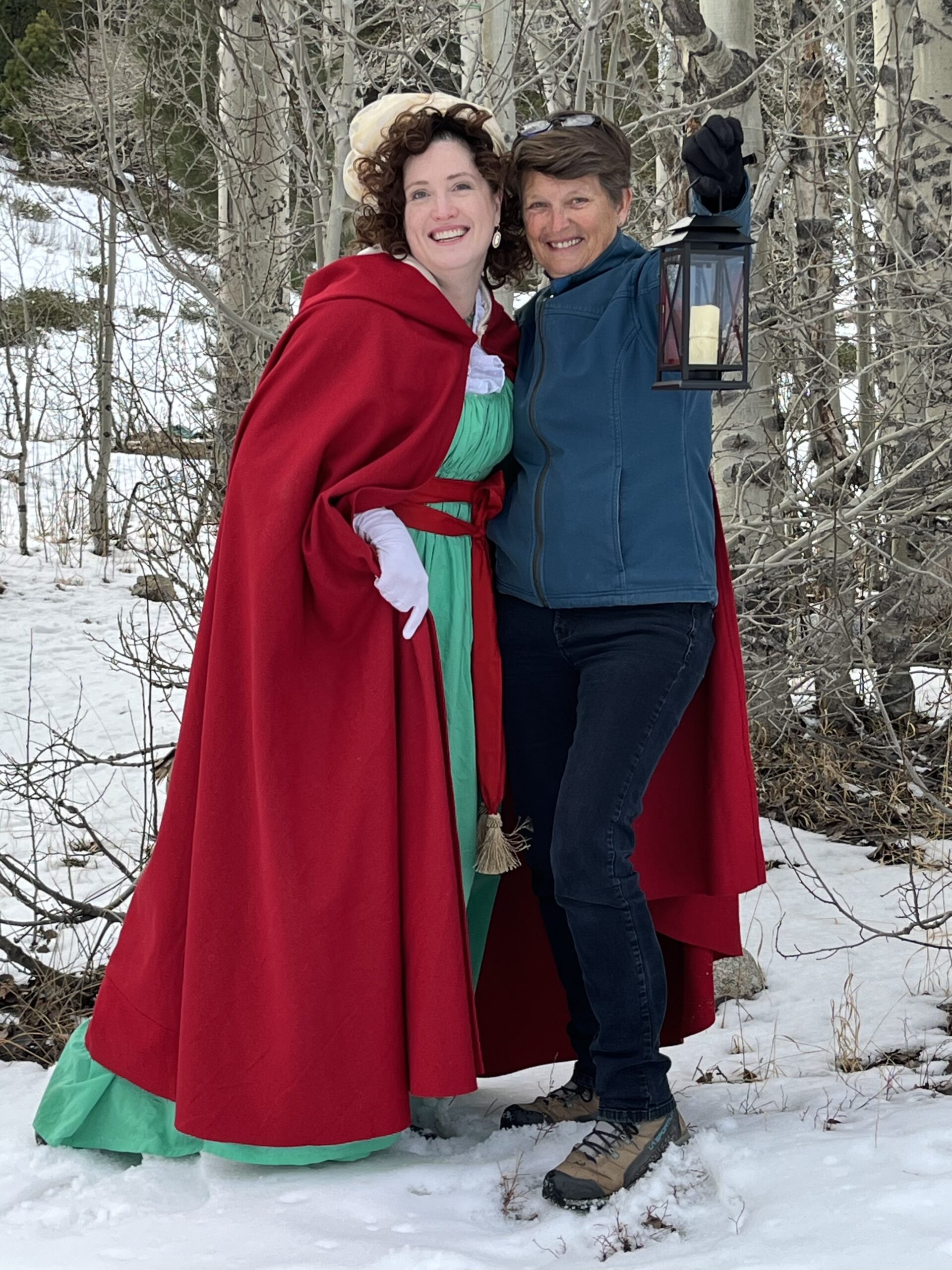 Tabubilgirl, wearing a long red cardinal cloak, stands with a woman wearing a modern blue snow jacket and black jeans. The woman is holding a lantern.