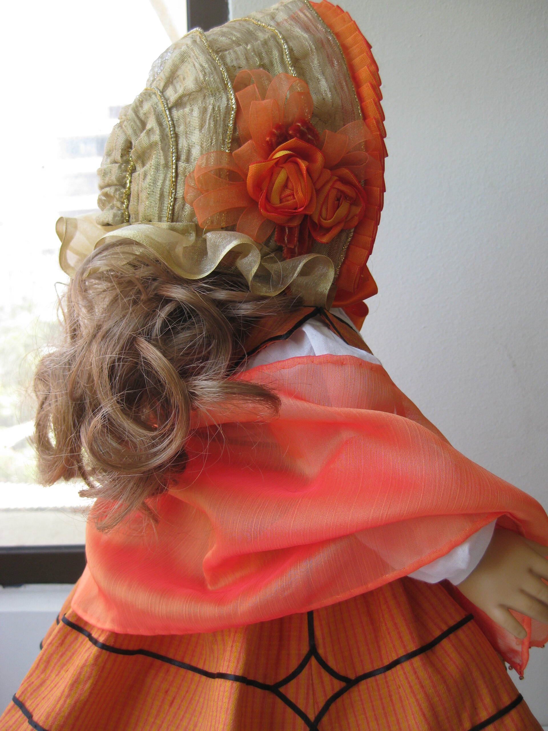 An American girl doll wears a drawn bonnet with an orange shawl and dress